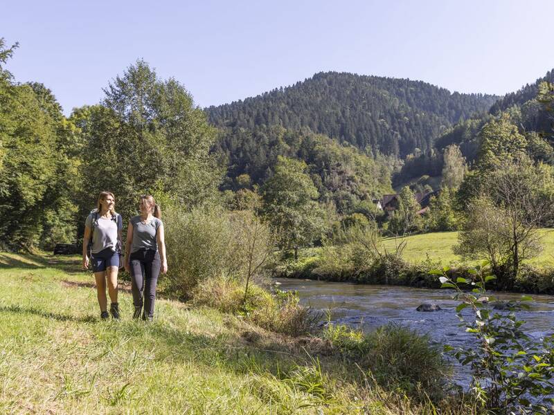 Zwei Wanderinnen laufen über eine Wiese am Ufer der Kinzig mit bewaldeten Hügeln im Hintergrund