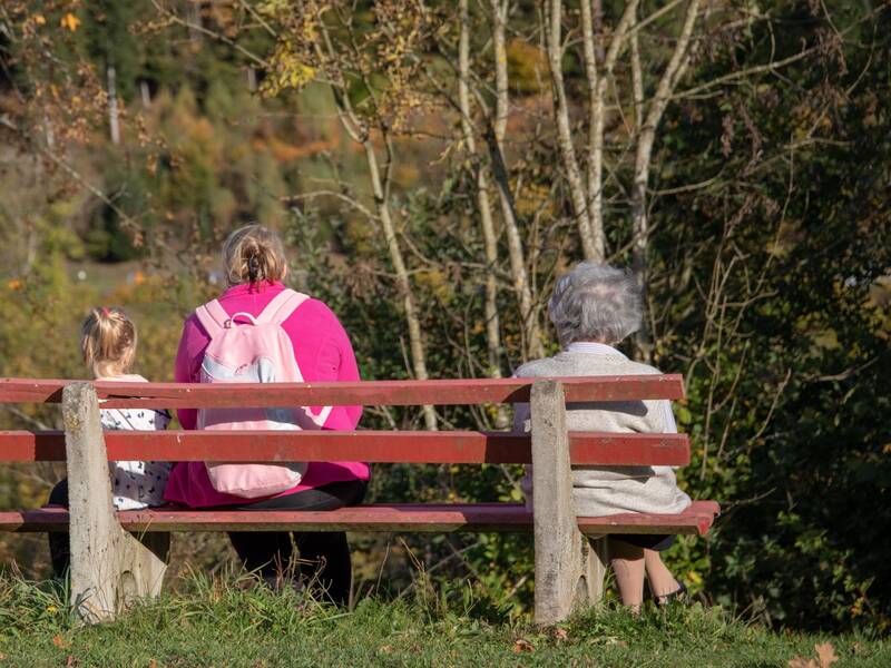Drei Personen unterschiedlichen Alters sitzen nebeneinander auf einer roten Bank und blicken auf eine herbstliche Landschaft mit Bäumen und einem Fluss.