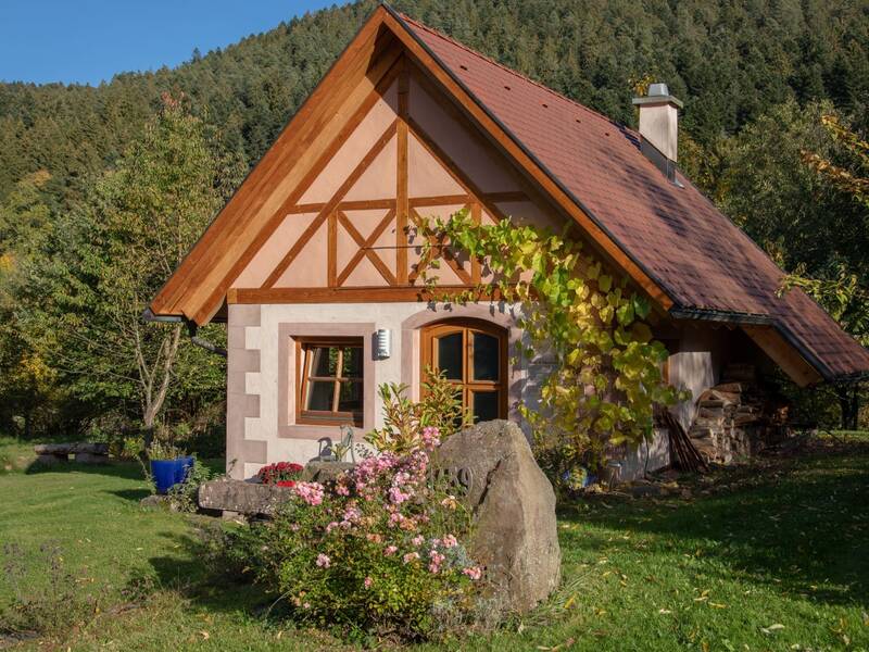 Ein kleines, gemütliches Häuschen mit Fachwerkdetails, umgeben von einem üppigen Garten und grünen Bäumen, steht im Vordergrund einer bewaldeten Hügellandschaft an einem sonnigen Tag.