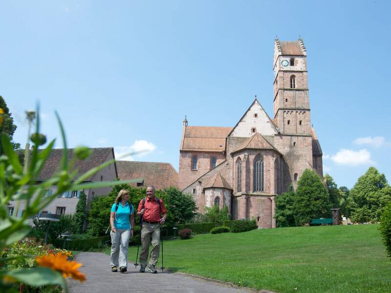 Ein Wanderpaar geht durch einen Park vor dem Alpirsbacher Kloster unter blauem Himmel.