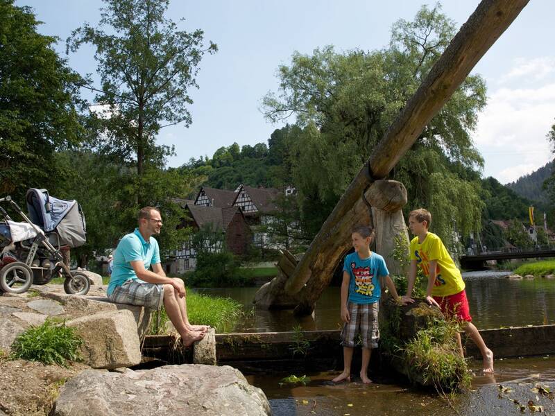 Eine Familie verbringt Zeit am Ufer der Kinzig in Schiltach; ein Mann sitzt auf einem Stein, neben ihm spielen zwei Kinder im Wasser, während eine Frau mit einem Kinderwagen neben ihm steht.