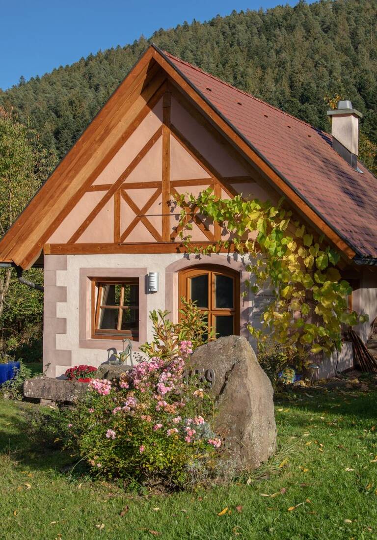 Ein kleines, gemütliches Häuschen mit Fachwerkdetails, umgeben von einem üppigen Garten und grünen Bäumen, steht im Vordergrund einer bewaldeten Hügellandschaft an einem sonnigen Tag.