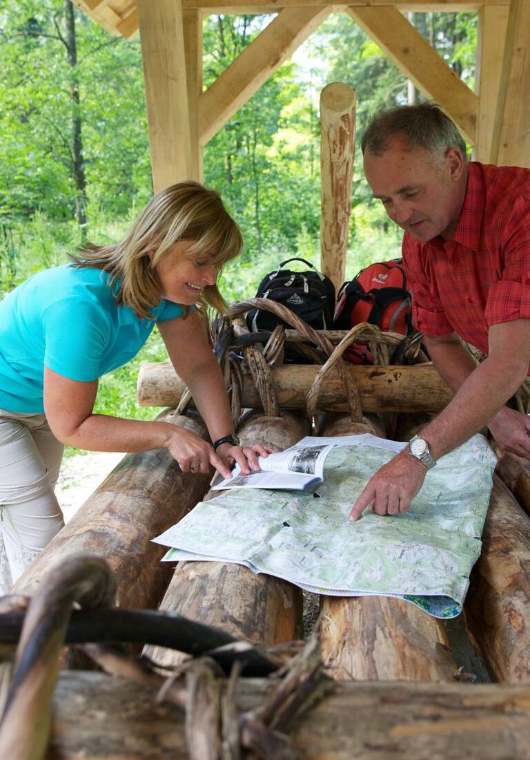 Ein Mann und eine Frau stehen an einem originalgetreuen Schaufloß unter einem Holzdach im Wald und schauen auf einer Landkarte nach dem Weg,.