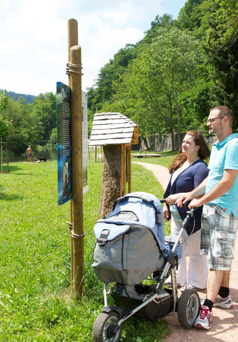 Zwei Personen stehen vor einer Informationstafel des Flößerpfads in einer grünen Parklandschaft und lesen die darauf befindlichen Informationen.