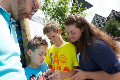 Ein Erwachsener und drei Kinder betrachten gemeinsam etwas auf einem Smartphone im Freien.